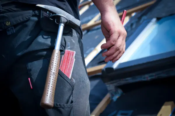 Dachdeckermeister mit Hammer und Messstab an seinem Gürtel steht auf einer Leiter, bereit, Reparaturen am Dach durchzuführen.